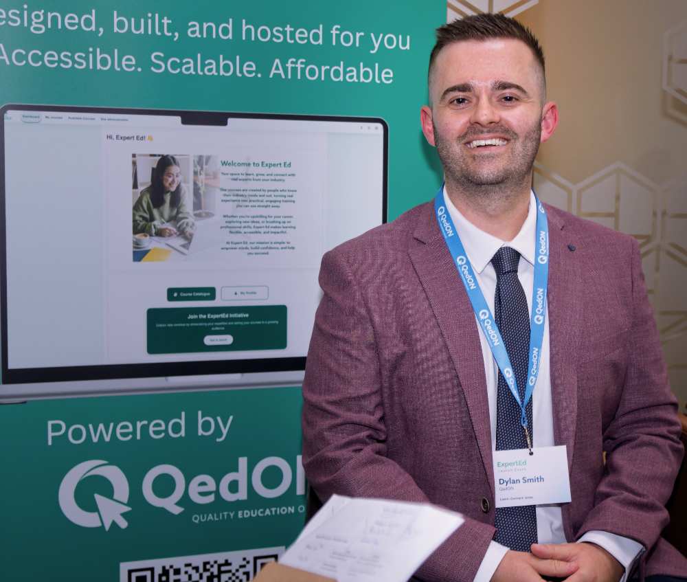 Individual standing in front of an ExpertEd promotional display at an event, wearing a lanyard and badge, with a screen showing the platform interface and text describing accessible, scalable and affordable eLearning powered by QedON visible in the background.