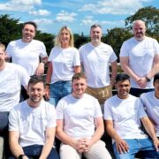 Team photo of TensorX staff outdoors on a rooftop terrace, with ten people wearing white company T-shirts, seated and standing in two rows against a backdrop of trees and blue sky.