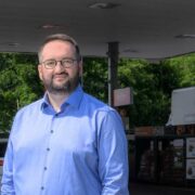 erson wearing a blue shirt and glasses standing at a fuel station forecourt, with petrol pumps, signage, a parked car, and a convenience store visible in the background under a canopy structure.