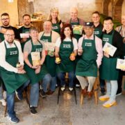 Group of food entrepreneurs wearing green Grow with ALDI aprons posing indoors in a rustic setting, each holding packaged food products including sauces, snacks, baked goods and ready meals from their brands.