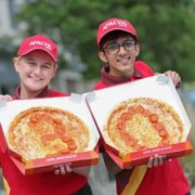 Two Apache Pizza staff members in red uniforms and branded caps hold open pizza boxes displaying pizzas arranged with pepperoni slices forming the number “30,” marking the chain’s 30th anniversary, with an outdoor urban background softly blurred behind them.