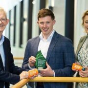 Three people stand indoors along a railing in a modern building, holding small orange containers and a green glass award. The central person holds a triangular green plaque with visible text reading Student Entrepreneur Awards, while the two people on either side present AURA-branded sports cream tubes. The background shows a bright corridor with large windows and soft natural light.