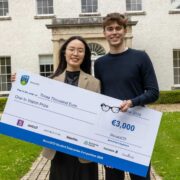 Two people stand outdoors in front of a large, light-coloured building with tall windows and a central doorway, holding an oversized cheque. The cheque displays the UCD and NovaUCD logos and reads “One to Watch Prize” with a value of €3,000, dated June 2026. Both individuals face the camera and hold the cheque together, with a grassy lawn and pathway visible in the foreground.