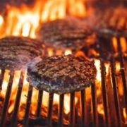 Close-up of beef burgers grilling on a barbecue, with flames rising through metal grates and charring the patties over an open flame.