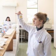 Laboratory technicians in white lab coats examine liquid samples in a bright, clean testing facility, with one technician holding up a small vial while others work with filtration equipment and measurement tools along a long workbench.