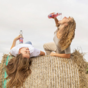 Two people are sitting on top of a large round hay bale in an open rural field. One person lies back across the hay bale holding a bottle upright, while another person kneels and drinks from a similar bottle tilted upward.