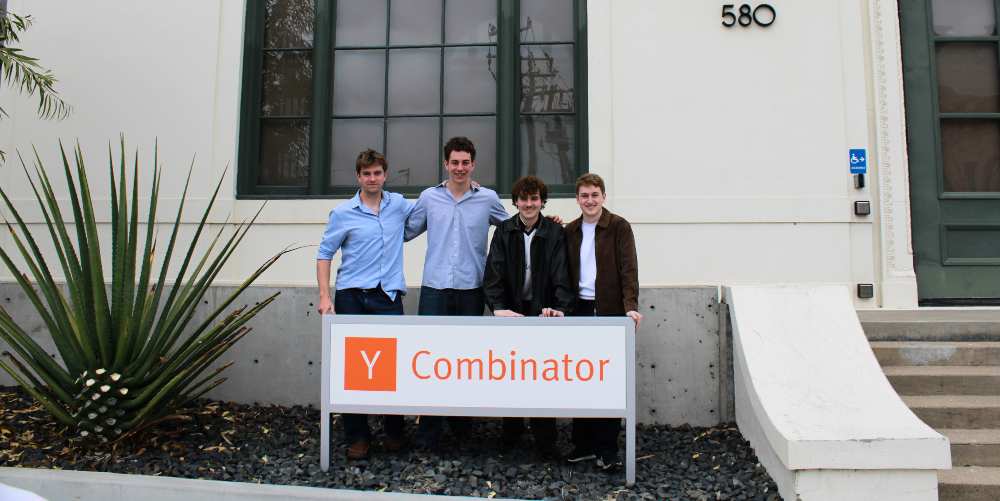 Four founders stand outside a building behind a sign reading “Y Combinator,” posing together in front of large windows and a numbered entrance.