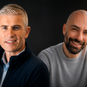 Two business professionals photographed side by side against a dark background, one wearing a dark jacket and collared shirt and the other in a light grey top, both facing the camera in a studio-style portrait setting.