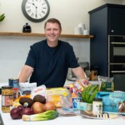 Person standing behind a kitchen counter with a variety of groceries spread out in front, including fresh vegetables, packaged bread, avocado, onions, chickpeas, and branded food products. The setting is a modern kitchen with dark cabinets, built-in ovens, a white countertop, a wall clock, and a wooden shelf holding a plant and small appliances in the background.