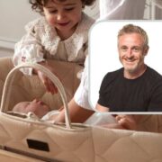 Baby lying in a cushioned Moses basket while a toddler reaches in, with a small inset headshot of a man overlayed on the right side of the image.