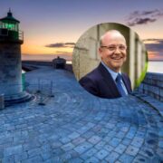 Circular image overlay of a suited speaker set against Dun Laoghaire harbour at sunset, featuring a stone pier and green lighthouse overlooking calm sea waters.