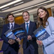 Three individuals standing in a modern office lobby hold copies of a printed report titled &ldquo;The hidden cost of cyber risk,&rdquo; with a reception desk and contemporary lighting visible in the background.