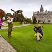 Falconry demonstration on the grounds of a large historic estate in Ireland, with a bird of prey in flight toward a handler while a small group watches and a photographer captures the scene, with landscaped lawns and a grand mansion in the background.