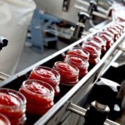 Glass jars filled with red sauce moving along an automated production line in a modern food manufacturing facility, with stainless steel machinery and conveyor system in view.