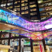 Glass pedestrian bridge connecting modern office buildings at night, illuminated with vibrant multicolour LED lighting, with lit office windows in the background in Dublin.
