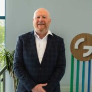 Person in a dark checkered suit and white shirt standing indoors beside a large window, with a wooden circular sign displaying a stylised &ldquo;G&rdquo; logo mounted on vertical coloured strips, and a potted plant visible in the background.