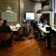 Group of people working on laptops during a startup hackathon in a modern office, listening to a presentation displayed on a screen in a collaborative workspace.