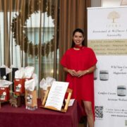 Display stand showcasing IPERA skincare products at an indoor event, with a branded banner reading &ldquo;IPERA Hormonal & Wellness Support Naturally,&rdquo; a table of packaged items and gift boxes, and a person in a red outfit standing beside the display with a decorative wreath and curtains in the background.
