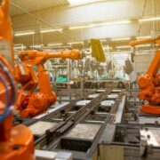 Industrial robotic arms working on an automated assembly line inside a modern manufacturing facility, with orange machines positioned along metal conveyor tracks under bright factory lighting and safety enclosures visible in the background.