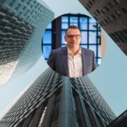 Portrait of a business professional overlaid on a low-angle view of modern glass office buildings, symbolising corporate finance, fintech innovation and global growth.