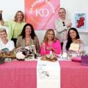 Group of six people seated and standing behind a display table of Irish-made products at the Kilkenny Design Makers Academy launch, with a pink Kilkenny Design KD Makers Academy backdrop. The table features candles, skincare jars, mugs, textiles, a small sheep craft item, books, and handbags, while one person holds up a small black shopping bag and another presents a framed map artwork.