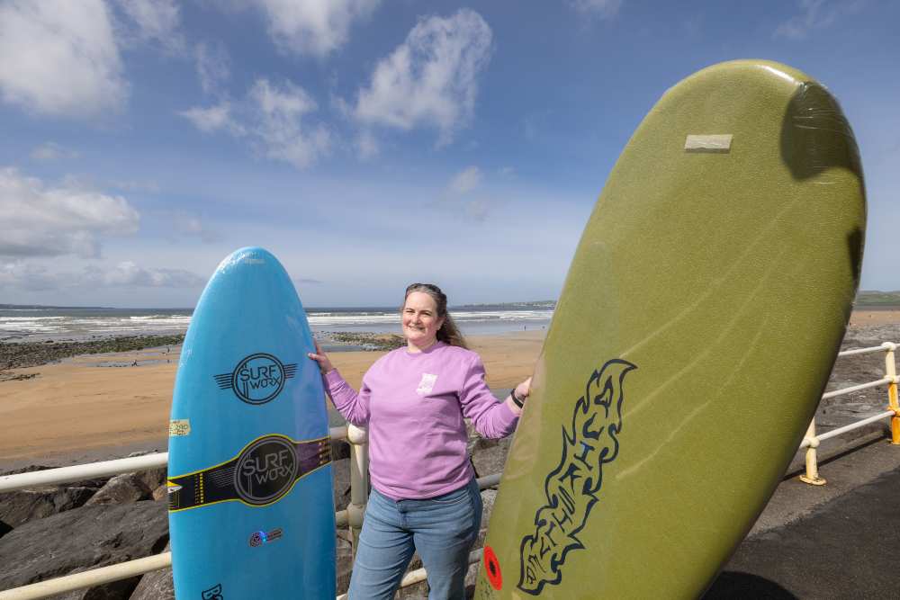 Person standing on a seaside promenade in Lahinch with two upright surfboards—one blue with “Surf Worx” branding and one green with a flame-style logo—set against a sandy beach, rocky shoreline, and ocean waves under a partly cloudy sky.
