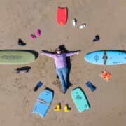 Overhead view of a person lying on a sandy beach surrounded by neatly arranged surfing gear, including two surfboards, bodyboards, flippers, wetsuit boots, and other accessories positioned in a circular layout.