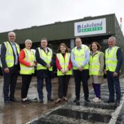Group of seven people wearing high-visibility vests standing outside a Lakeland Dairies processing facility in Ballyrashane, with a branded building and delivery truck visible in the background on a wet day.