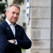 Professional portrait of a person in a navy suit with arms crossed, standing outdoors beside a stone building, with greenery and a blurred urban background in Dublin.