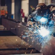 Welder working on a metal beam in an industrial workshop, wearing protective gear and helmet, with bright sparks flying from active welding equipment and machinery visible in the background.