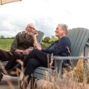 Two older adults sitting in outdoor lounge chairs in a garden setting, enjoying drinks under a parasol with a green countryside landscape in the background.