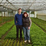 Two people standing together inside a greenhouse, surrounded by rows of leafy green crops, with metal framing and a curved transparent roof structure extending into the background.