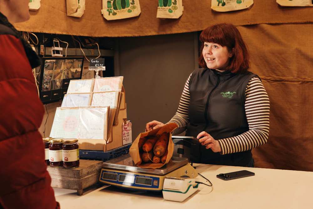 Person serving a customer at a market stall, placing carrots in a paper bag on a weighing scale. The counter also displays jars of preserves, greeting cards with a small sign reading “Cards €3.95,” and a payment terminal, with packaged goods hanging in the background.