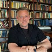 Person seated at a wooden table in a room lined with bookshelves filled with books, wearing a black shirt and wristwatch, with hands resting on the table in a study or library setting.