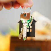 Close-up of a hand holding a set of house keys attached to a keyring with a small house-shaped keychain, with a blurred model of a home in the background.