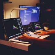 Person working at a desk with a laptop and external monitor displaying code, alongside a tablet, desk lamp and plant in a dimly lit workspace, illustrating digital work and AI or software development context.