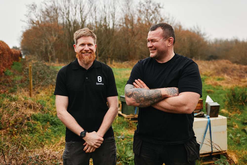 Two people standing outdoors in a rural, grassy area with trees in the background, beside beekeeping equipment including hives and boxes. One person wears a black polo shirt with an “Openhive” logo, while the other wears a plain black T-shirt and has visible arm tattoos.