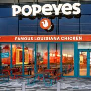 Popeyes fast food restaurant exterior with large illuminated signage and orange branding, glass-fronted entrance, and outdoor seating area with picnic tables at a modern roadside location.