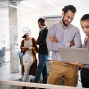 Two professionals standing in a modern open-plan office reviewing a laptop together, with colleagues working in the background, representing workplace collaboration and digital upskilling.