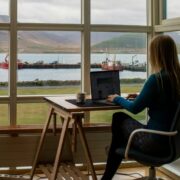 Person working on a laptop at a desk by a large window overlooking a harbour, with boats, mountains and water visible outside in natural daylight.