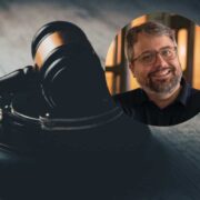 Close-up of a wooden judge&rsquo;s gavel resting on a desk, symbolising the legal system, with a circular inset showing a person wearing glasses against a softly lit indoor background in the upper right corner.