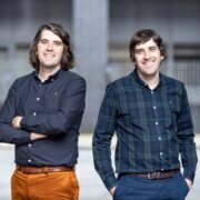 Two adults standing side by side outdoors in an urban setting, both wearing button-down shirts and trousers, with a blurred modern building fa&ccedil;ade in the background. One adult stands with arms crossed in a navy shirt and brown trousers, while the other has hands in pockets wearing a dark green check shirt and blue trousers.