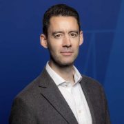 Professional corporate headshot of a business executive wearing a grey blazer and white shirt against a blue background, representing leadership, technology, business strategy and corporate innovation.