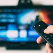Hand holding a TV streaming remote control in front of a smart television displaying a video-on-demand entertainment platform, illustrating growth in Ireland’s digital media, streaming and connected entertainment market.