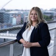 Business professional standing on a balcony overlooking Dublin&rsquo;s docklands, with the Samuel Beckett Bridge in the background.