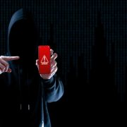 SEO-friendly alt text: Hooded figure holding a smartphone displaying a cyber attack warning against a dark digital background, illustrating cybersecurity risk.