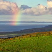 Rainbow over Ireland’s Atlantic coastline, with green farmland, rolling hills and a grazing cow overlooking the sea.