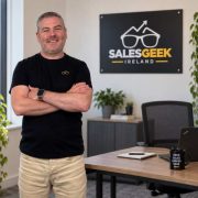 Jonathan Barker standing in the Sales Geek Ireland office beside a desk and company branding, as the sales leadership consultancy expands support for Irish SMEs and business growth.