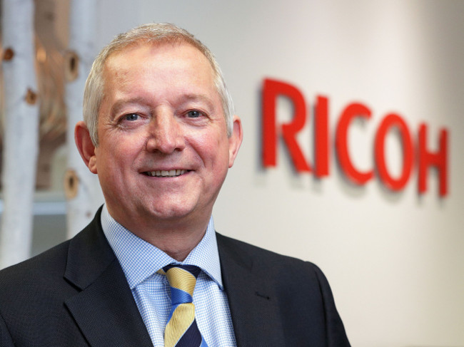 Man in dark suit with blue shirt and tie standing in front of a wall with words Ricoh on it.