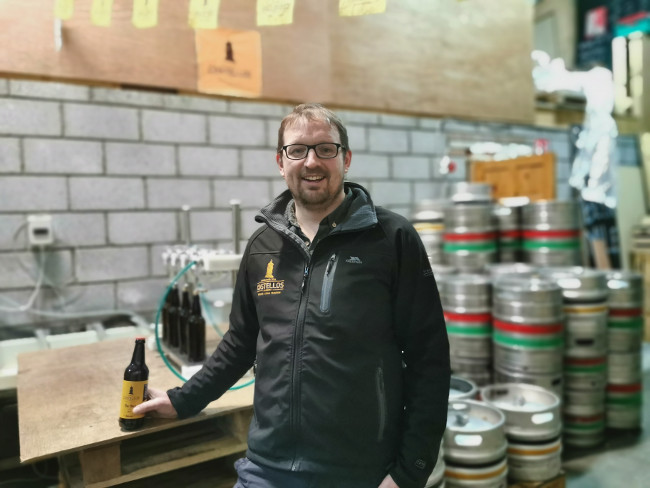 Man standing with a bottle of beer in front of beer kegs.